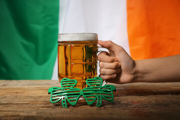 Female hand with mug of beer and plastic eyeglasses on wooden table against flag of Ireland. St....