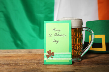Mug of beer with festive card and leprechaun hat on wooden table against flag of Ireland. St....