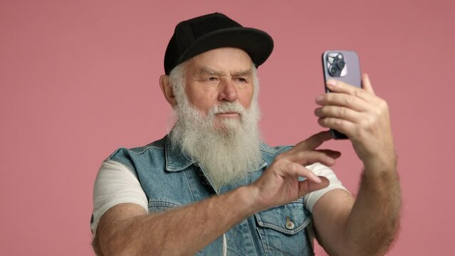 A playful elderly man with a white beard and cap is making a funny face while taking a selfie, showing his humorous side on a pink background. Camera 8K RAW. 