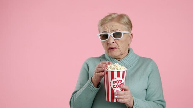 Terrified senior woman eating popcorn from paper cup, standing over pink background, lowering her glasses, closing eyes with hand being terribly frightened. High quality 4k footage