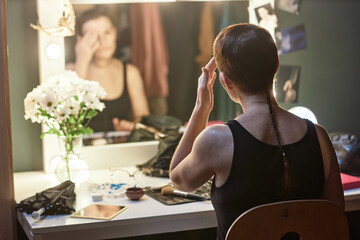 Back view of female artist doing make up by vanity mirror backstage in theater preparing for...