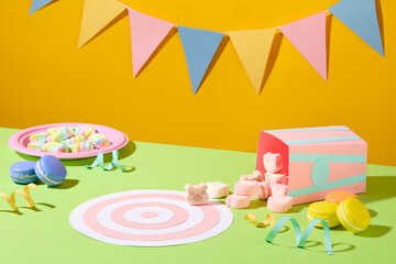 A decorative triangular flag string on a yellow background. Marshmallows, macarons and colorful ribbons are randomly arranged on a pastel green surface.