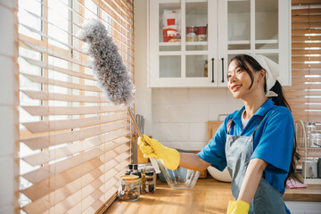 Enjoying housework Asian woman stands dusting window blinds. Smiling she holds a duster. Occupation...