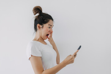 Asian Thai woman holding mobile phone with funny surprised and shocked face, while looking cellphone, standing isolated over white background wall.
