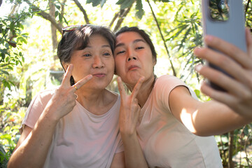 Asian Thai daughter and elder mother taking selfie together with happiness, showing hand two fingers gesture while spending time together at cafe in garden on holidays.