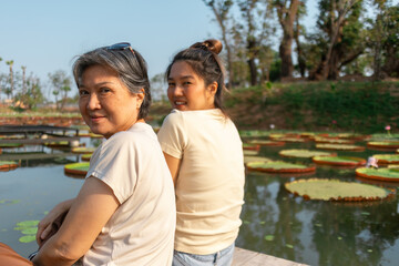 Backside view of asian Thai Chinese elder mother and daughter sitting on the bank by lotus flower lake local lagoon, both woman happy smiling, travel on vacation.