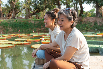 Side view of asian Thai Chinese elder mother and daughter sitting on the bank by lotus flower lake local lagoon, both woman happy smiling, travel on vacation.