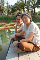 Side view of asian Thai Chinese elder mother and daughter sitting on the bank by lotus flower lake local lagoon, both woman happy smiling to camera, traveling on vacation.