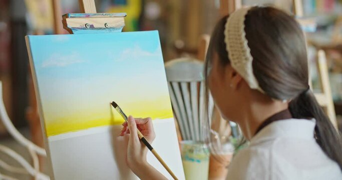 Back view of Attractive beautiful asian stylish female artist painting picture in art studio. Young woman painter draws with paint brush and oil colors creating artwork sunset landscape on canvas.
