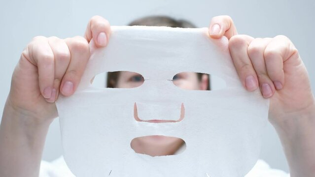 Portrait of a young woman in a white bathrobe holding a sheet moisturizing mask front view close-up.