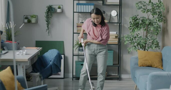 Young Asian housewife washing floor in apartment using modern mop focused on housework at home. Domestic chores and lifestyle concept.