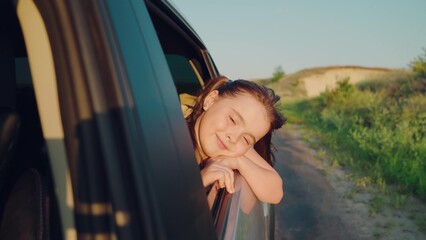 child girl face looks smiling from car out window, bright light setting sun through his fingers,...