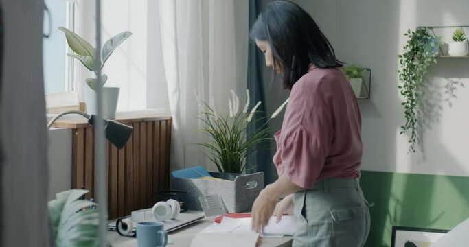 Cheerful Asian lady tidying desk putting away papers and stationery concentrated on clean-up in apartment. Lifestyle and housework concept.