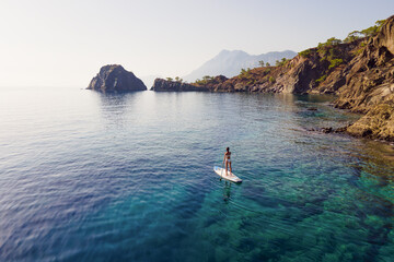 Woman in bikini stand on sup paddle board with paddleboarding passing calm sea with rocky shore....