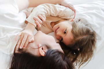 Portrait of attractive smiling young mother and little daughter lying on bed on white bed linen