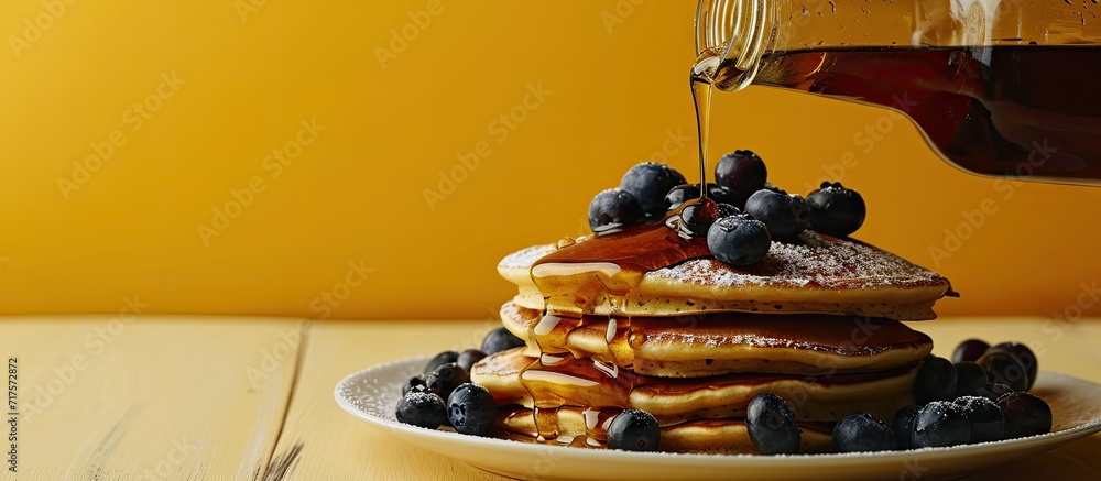 Poster horizontal image of maple syrup pouring onto pancakes and blueberries on yellow with copy space food