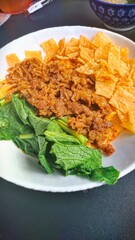a portion of biang-biang noodles. The noodles are wide and served with a distinctive red sauce, topped with vegetables, wonton chips and pieces of beef or chicken