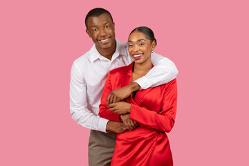Affectionate black couple posing, man in white, woman in red, pink backdrop