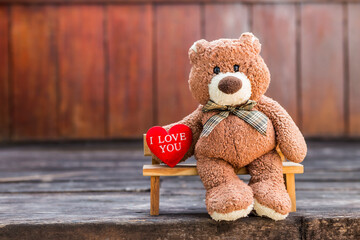 Brown teddy bear with I love you red heart on wooden bench with space on blurred wood background,...