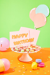 The words HAPPY BIRTHDAY on a pink card, gummy candy on a plate. Balloons, macarons and confetti decorate an orange surface with a pastel green background.