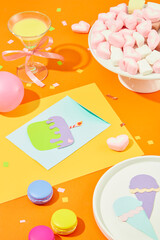 A plate of marshmallows, a cocktail, Ice cream and a birthday cake cut from colored paper, balloons and macarons on an orange background.