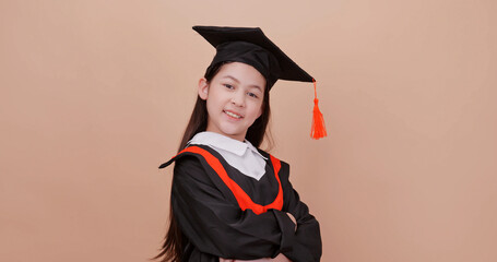 A school girl wearing a graduation gown makes happy gestures and expressions. Isolated on brown background in studio.