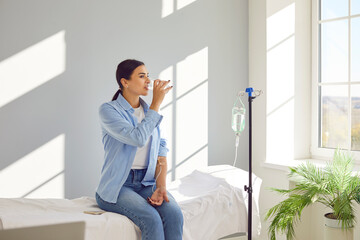 Woman with sterile tube in arm sitting by pole with IV bottle at medical center, drinking water and receiving vitamin therapy infusion to boost body immune system, preserve beauty or fight hangover - obrazy, fototapety, plakaty