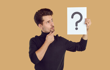 Pensive man is looking at piece of paper with question mark drawn on it, which he is holding in his hand. Young Caucasian man holding question mark symbol and thinking isolated on beige background. - Powered by Adobe