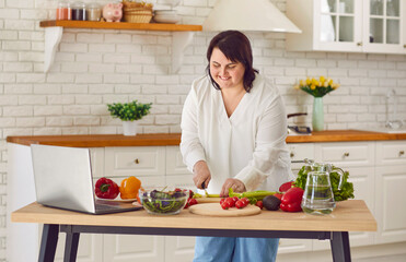 Portrait of fat overweight young woman preparing fresh vegetable salad looking at recipes on a...