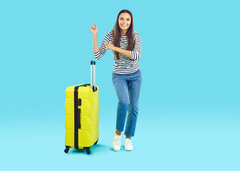 Full length portrait of happy woman standing with suitcase. Beautiful casually dressed young woman with luggage having cheerful trip dancing over isolated on light blue background