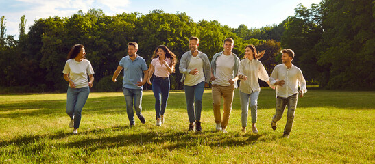 Cheerful young men and women having fun running on green lawn in park on sunny summer day. Concept of people and friendship. Friends in comfortable casual clothes having fun outdoors. Panoramic banner - obrazy, fototapety, plakaty