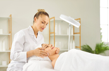 Happy pretty young woman lying on a massage bed under a magnifying lamp and enjoying a face massage...