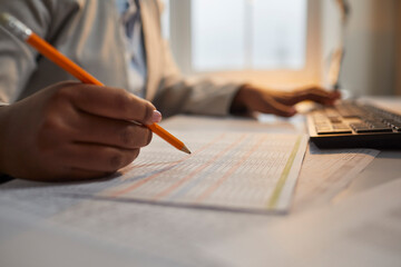Fototapeta premium Professional accountant working in office, doing business paperwork, checking some data. Close up of hand of African American woman holding pencil over paper data sheet, with computer in background