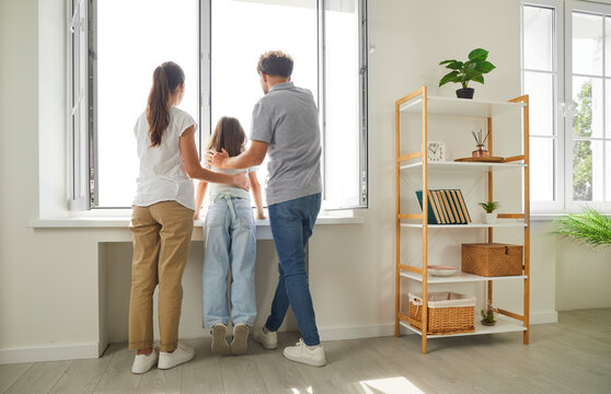 Happy Young Family And Their Preteen Daughter Are Enjoying View In Their New Apartment. Rear View Of Family With Child Looking Through Window In Living Room. Moving And Family Togetherness Concept.