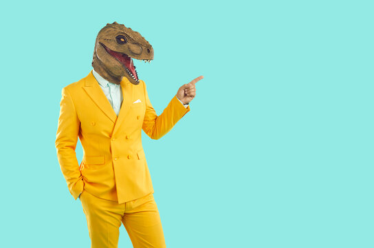 Stylish man in funny dinosaur mask, points to an empty space to copy. Funny showman in bright yellow suit and white shirt on an isolated pale blue background.