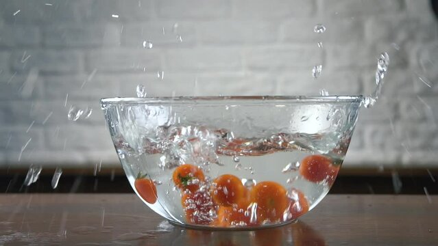 Fresh organic red cherry tomatoes with green tails fall into a glass transparent bowl with water with big splashing and spraying on a wooden table on the background of a white brick wall. Slow motion