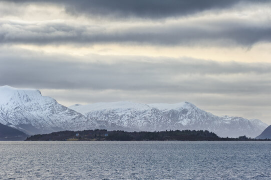 The serene image captures a majestic snow-covered mountain range looming above the calm, reflective ocean waters under a cloudy evening sky.