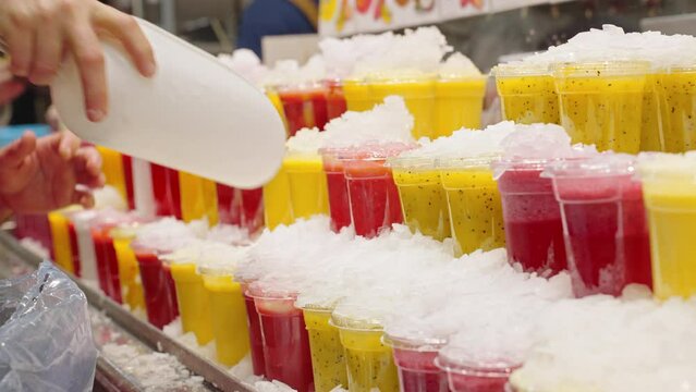 Showcase with cold freshly squeezed strawberry, mango juices in glass for takeaway Central market of Barcelona, Spain. Hand male merchant pours ice crush on products to preserve freshness. Healthy 
