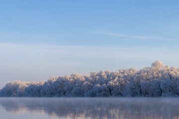Naklejka na ściany i meble A winter tableau unfolds along the river’s edge. Nature’s brush has painted a serene masterpiece — the water's surface a pristine canvas, adorned with a delicate quilt of snow.
