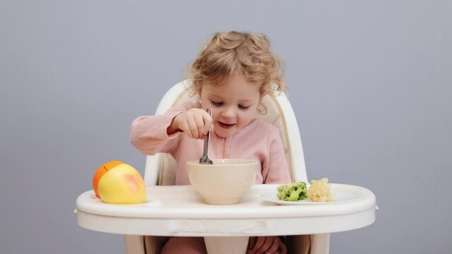 Baby and healthy nutrition. Breakfast for the little kid. Child's food. Taking care of child's health. Charming little baby girl with wavy hair eating in highchair isolated over gray background