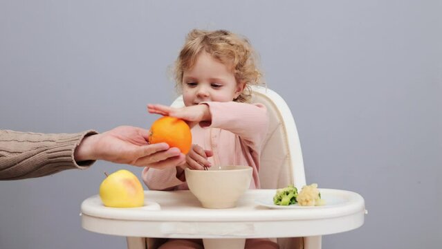 Funny little toddler blonde wavy haired girl eating by herself in highchair isolated over gray background unrecognizable mother giving fresh orange to kif for healthy dessert
