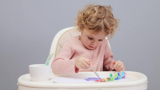 Funny curly haired toddler girl sitting in highchair painting with watercolors creating picture with multicolored paints playing alone isolated over gray background