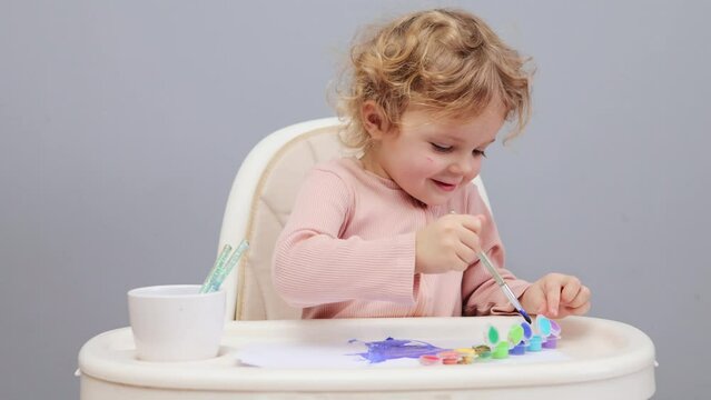 Creative kid. Education through color. Learning to draw. Child's daycare activities. Smiling joyful curly haired blond in highchair painting with watercolor isolated over gray background