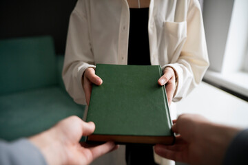 A close-up view captures the moment one person hands over a green hardcover book to another,...