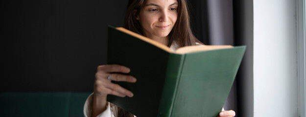 A woman stands by a large window, bathed in natural light, deeply focused on reading an open book.