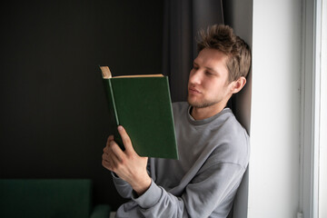 A focused young man wearing a casual grey sweater is deeply absorbed in reading a green hardcover...