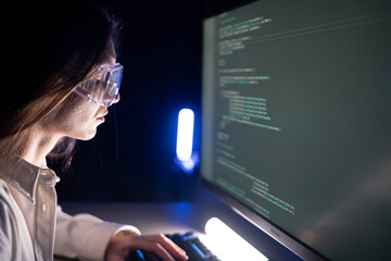 A focused woman works diligently on her coding assignment in front of a computer screen in a dimly lit office after hours.
