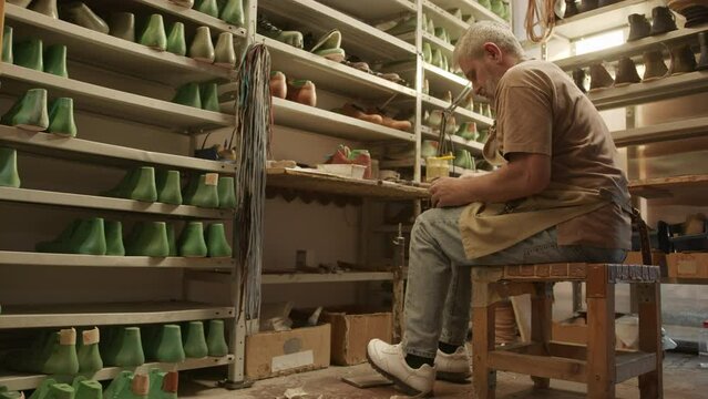 Full shot with copy space of mature shoemaker working in cobbler workshop with shoe lasts and supplies on shelves