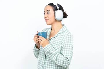 Beautiful young woman wearing green plaid pyjama and holding a cup wears stereo headphones listening to music concentrated and looking aside with interest.