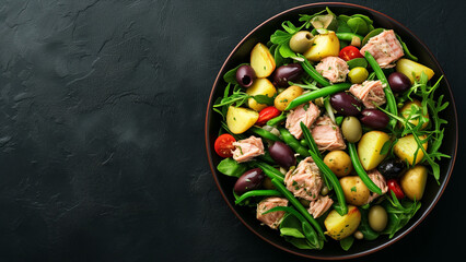 Top down view on a plate with a tuna salad with potatos and green vegetables on a black texture background with space for text